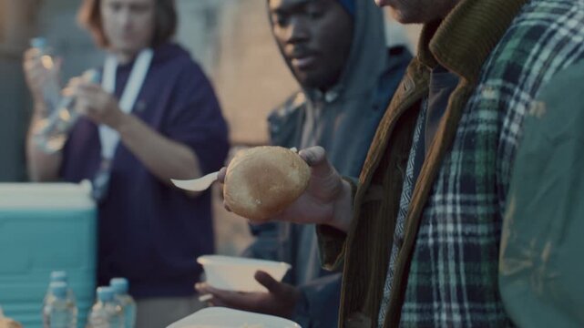 Group of volunteers providing water, bread, beans and pasta to diverse homeless men at food station during community outreach event in city alley