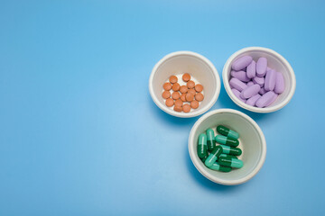 Three white bowls containing different types of medicine: round brown tablets, purple oval pills, and green capsules isolated on a blue background. Ideal for medical, pharmaceutical, and wellness