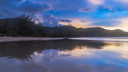 Evening after sunset on a tropical island. The clouds in the blue sky are highlighted in pink and gold. Silhouettes of mountains. Reflection on the smooth shiny surface of the ocean. Malaysia. Borneo.