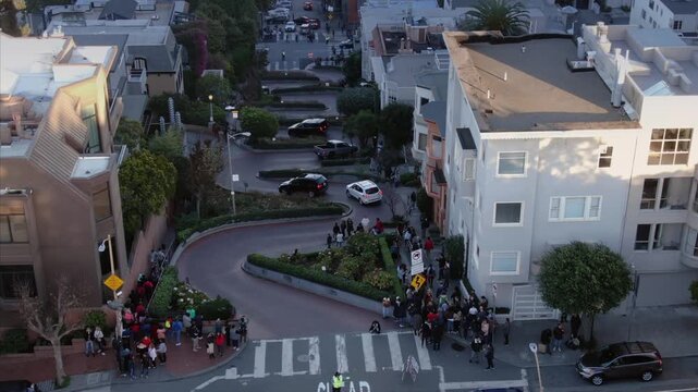 Lombard Street in San Francisco, USA, is a popular tourist destination. People are visiting and taking photos of the winding road and flower beds. A traffic controller is present.