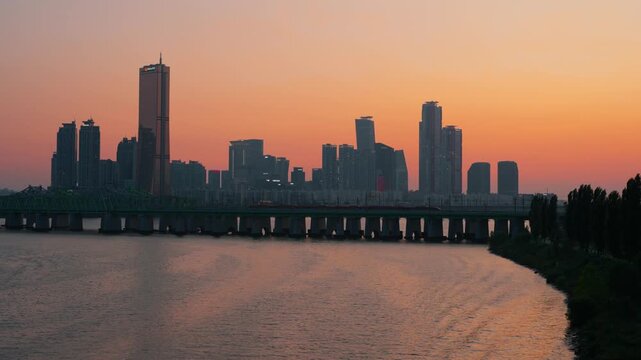 A beautiful sunset casts a warm orange glow over the Yeouido skyline and Hangang River in Seoul, South Korea, as a subway train crosses a railway bridge.