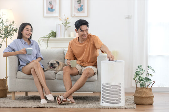young asian couple drinking morning coffee sitting on sofa relaxing time living room with small pug dog rests beside, man pressing power button air purifier for clean air in house for good health.