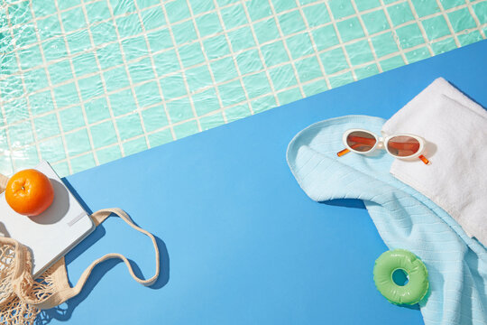 Bright summer setup with blue tiles, white towel, green float, and orange fruit near turquoise water, with soft space for product or design.