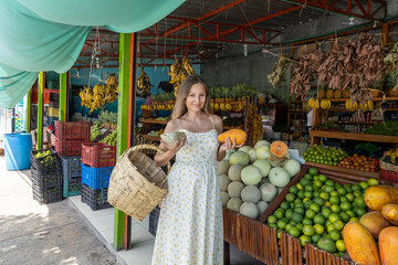 Pregnant woman weared in a light floral dress is shopping for fresh organic tropical fruits at a...