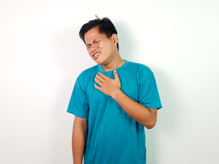 A young Asian man wearing a blue shirt expresses chest discomfort, isolated on a white background....