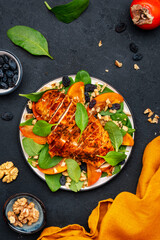Autumn persimmon salad with grilled chicken breast, walnuts, raisin and spinach, black background, top view