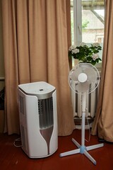 Air conditioner and standing fan beside a window with a potted plant and beige curtains