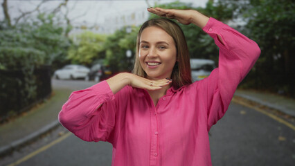 Fototapeta premium Woman smiling in pink shirt posing playfully outdoors on a city street with trees and cars in the background, exuding happiness and youthful energy.