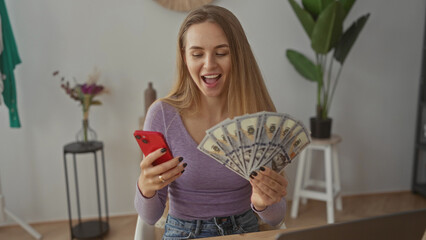 Young woman holding dollars and smartphone in modern living room, expressing joy, suggesting...