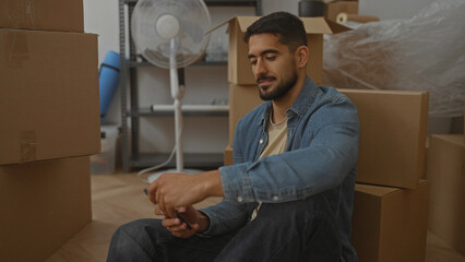 Young man sitting in new home surrounded by boxes, smiling while using phone in spacious living...