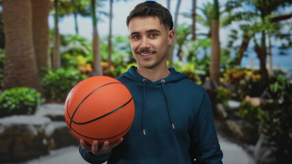 Young hispanic man holding a basketball in a sunny outdoor park showing a relaxed expression amidst lush greenery under clear skies.