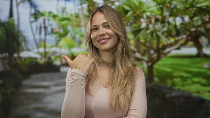 Naklejka premium Blonde woman pointing thumb to side on a sunny park walkway with tropical trees; direction confidence.