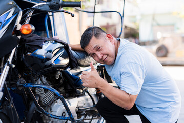 Middle-aged Southeast Asian tricycle driver kneeling fixing motorcycle engine, wearing blue shirt,...