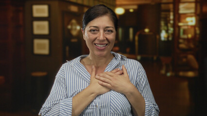 Fototapeta premium Middle age woman in striped shirt places hands on chest in building entrance under warm light; gratitude.