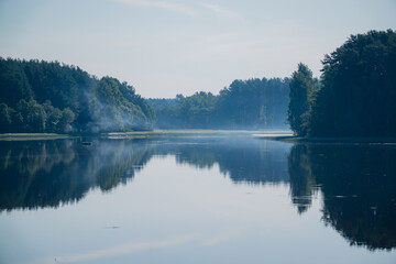 Fototapeta premium A gentle mist hovers over calm water, reflecting trees and creating a serene dawn
