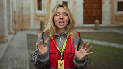 Fototapeta premium Young woman volunteer gesturing energetically in front of an old university building outdoors wearing a red vest and lanyard indicating her role in the community project setting.