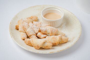 A plate overflows with sweet pastry rolls, lightly dusted with sugar, paired with a creamy dip