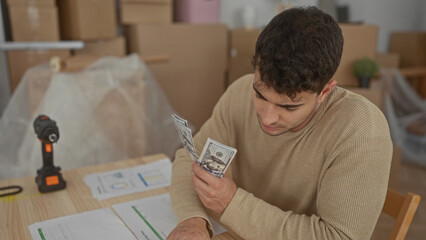 Man stacks bills then counts totals at home while a young hispanic tracks cash near new boxes on a...