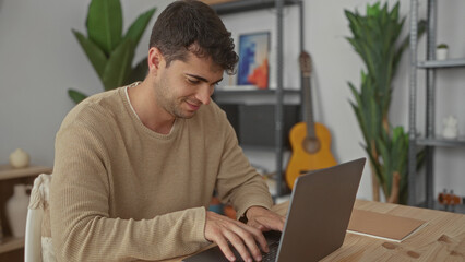 Man adjusts laptop and starts typing in home office as a hispanic young male computer freelancer, smiling contentedly.