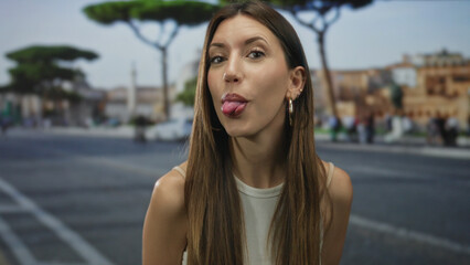 Young woman in white tank top with long brown hair sticks tongue out on urban street; cheeky...