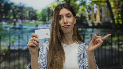 Young hispanic woman holds blank card with finger pointing near pond in forest; financial...
