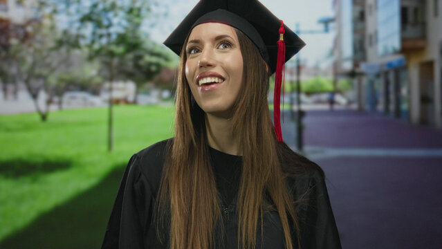 Profile young woman in mortarboard cap with red tassel and gown smiles broadly on city street; pride. - Powered by Adobe
