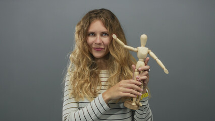 Woman holds mannequin while smiling in studio with blonde young hair framing a wooden posing figure and adjusting its casual posture.