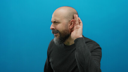 Obraz premium Bald man with beard gestures listening with hand to ear against vivid blue background.