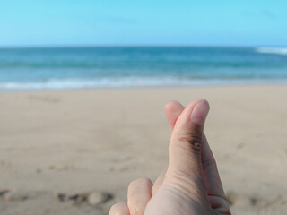 A close-up of a hand making a mini heart gesture against a blurred backdrop of a sunny beach and calm turquoise sea. Concept for love, vacation, or K-pop culture.