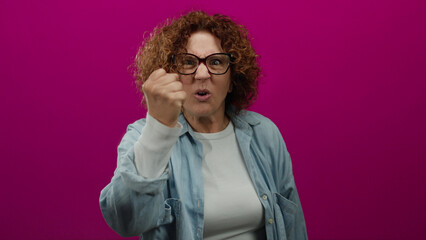 Middle age woman expressing anger with raised fist against vibrant pink background, wearing glasses and denim shirt, conveys emotion of frustration and determination.