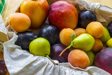 A beautifully arranged basket filled with a colorful mix of fresh fruits including red apples, green pears, dark plums, orange apricots, and nectarines. The basket is lined with a textured natural