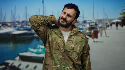 Hispanic man in military uniform with beard stands at a bustling port with boats visible, appearing to massage his neck under bright blue skies.
