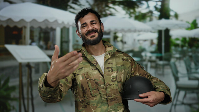 Hispanic man in military uniform with beard smiling holding helmet in restaurant outdoor seating area surrounded by tables and chairs under canopies.
