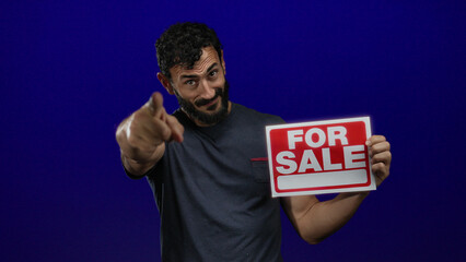 Hispanic man with beard holding a for sale sign against a blue background, pointing confidently...