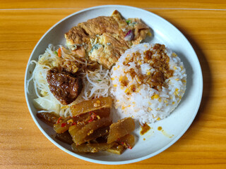 A typical Indonesian mixed rice dish with white rice sprinkled with spices, omelet, vermicelli, boiled vegetables and other side dishes on a white plate. Wooden table background.