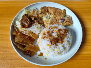 A typical Indonesian mixed rice dish with white rice sprinkled with spices, omelet, vermicelli, boiled vegetables and other side dishes on a white plate. Wooden table background.