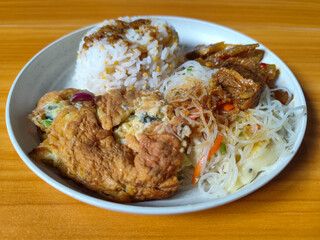 A typical Indonesian mixed rice dish with white rice sprinkled with spices, omelet, vermicelli, boiled vegetables and other side dishes on a white plate. Wooden table background.