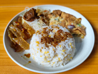 A typical Indonesian mixed rice dish with white rice sprinkled with spices, omelet, vermicelli, boiled vegetables and other side dishes on a white plate. Wooden table background.