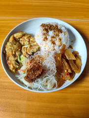 A typical Indonesian mixed rice dish with white rice sprinkled with spices, omelet, vermicelli, boiled vegetables and other side dishes on a white plate. Wooden table background.