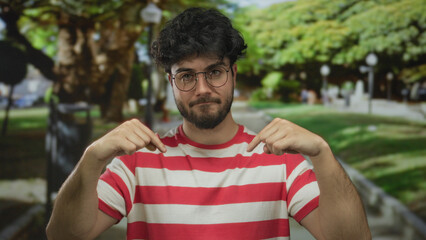 Young hispanic man with beard and glasses points downward while standing in a sunny park, wearing a...