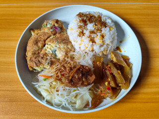 A typical Indonesian mixed rice dish with white rice sprinkled with spices, omelet, vermicelli, boiled vegetables and other side dishes on a white plate. Wooden table background.
