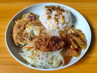 A typical Indonesian mixed rice dish with white rice sprinkled with spices, omelet, vermicelli, boiled vegetables and other side dishes on a white plate. Wooden table background.