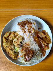 A typical Indonesian mixed rice dish with white rice sprinkled with spices, omelet, vermicelli, boiled vegetables and other side dishes on a white plate. Wooden table background.