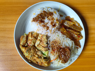A typical Indonesian mixed rice dish with white rice sprinkled with spices, omelet, vermicelli, boiled vegetables and other side dishes on a white plate. Wooden table background.
