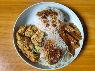 A typical Indonesian mixed rice dish with white rice sprinkled with spices, omelet, vermicelli, boiled vegetables and other side dishes on a white plate. Wooden table background.