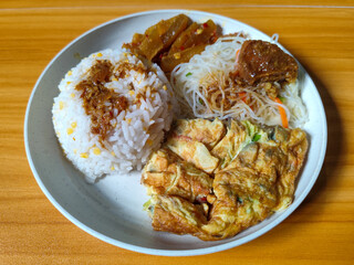 A typical Indonesian mixed rice dish with white rice sprinkled with spices, omelet, vermicelli, boiled vegetables and other side dishes on a white plate. Wooden table background.