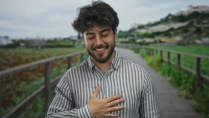 Young man with beard stands outdoors in a green park smiling and touching his chest serenely, enjoying the peaceful natural surroundings under a cloudy sky.