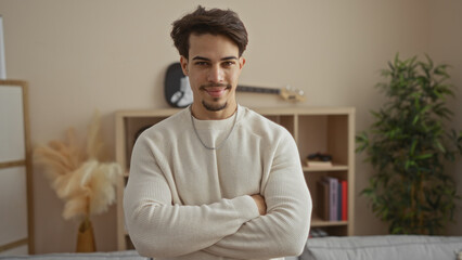 Fototapeta premium Young man with crossed arms standing confidently in a cozy living room, exuding a sense of calm and composure, surrounded by home decor elements in a modern apartment.