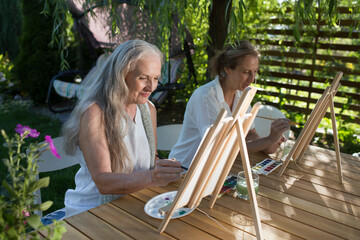 Two happy middle aged woman smiling while drawing watercolor as a recreational activity or therapy outdoors together with the group of retired women. KnowledgeForAll
