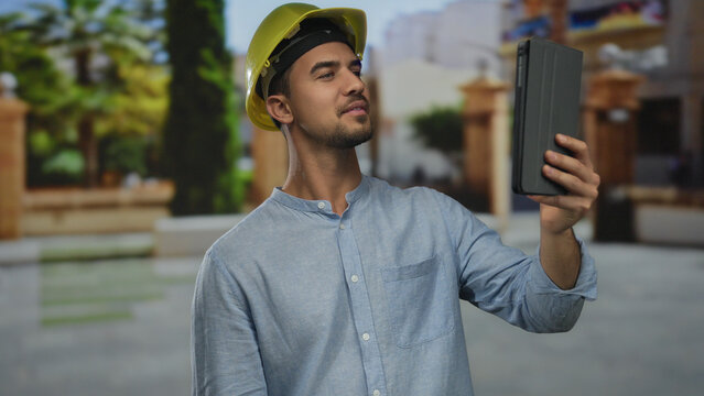 Young hispanic man with hardhat using tablet in outdoor park setting, demonstrating modern technology use in construction environment with greenery backdrop.